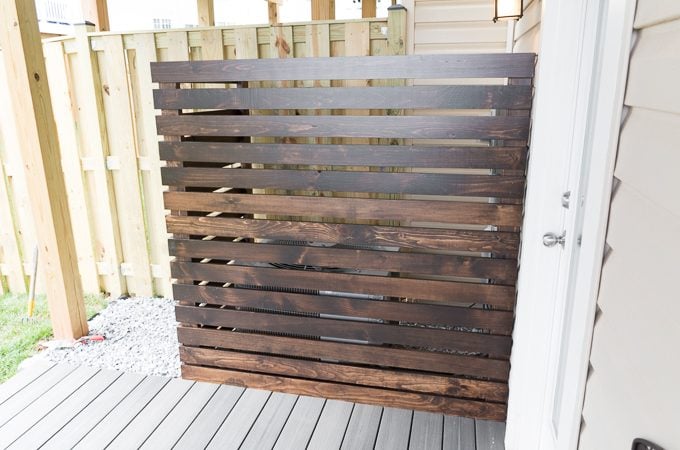 Hi guys, it's week 3 of the One Room Challenge, and we're finishing up the AC/utilities corner part of the yard. Wahoo! Last week, I shared progress on the platform deck and some light rock landscaping. This week, I'm sharing the DIY AC unit screen I made. I'm going to share how I did it, but I'm not going to share a complete tutorial. It's unlikely that anyone (except my next door neighbor) has exactly the same layout and needs as we do. Here's what we used to make a DIY AC unit screen: (Affiliate links below. Thank you for your support!) 2x4s for the standing pieces 1x4s for the slats Random orbit sander Fine-frit sandpaper to polish by hand Saw, finish nailer Liquid Nails Heavy Duty Construction Adhesive Varathane wood stain in Kona Minwax Helmsman Spar Urethane in Satin Kreg Jig and 2.5" pocket hole screws And here's how we did it. (Remember to wear a mask and eye protection while sanding and working with wood, and wear an appropriate mask while working with stains and finishes. Follow the directions and warnings from your particular brand. Do not use any tools without proper training, precautions, and supervision. Read my full disclaimer here.) Step 1: First we measured how big we wanted the screen to be. We decided to shape it like an L and have it come up high enough to cover other utilities, not just the AC unit. (If you make a project like this, make sure it does not interfere with anything that requires air circulation or regular reading.) After deciding on measurements, and knowing we wanted each screen slat to be 1 inch apart, we calculated how much 1x4 we'd need. Once we had the final height (14 pieces of 1x4, each an inch apart), we cut the four pieces of 2x4 to height. (Yes, that's a diaper and a package of wipes in my purse. Times have changed.) Step 2: Next I gave each piece a quick sand using my random orbit sander. This was mostly to polish each piece up and break down any rough or splintery edges. After each piece was cleaned up, I wiped the whole lot down and stained each using the Varathane wood stain in Kona. I also drilled pocket holes in one of my 2x4s to create the main corner support (but didn't attach them yet). This, coupled with the next step, was by far the most time consuming. (But was faster using a small roller instead of a paint brush!) I was VERY GLAD at this point that I hadn't used 1x2 like I'd originally wanted. That would have been painful. Step 3: Once I had stained everything and it had dried according to my instructions, I gave each piece a few coats of the Minwax Helmsman Spar Urethane in Satin. Oof, talk about a slow process. (See my post on how to stain and finish wood here.) And one that took over my garage for several days. Luckily the urethane dries in about 4 hours, so that helped me get two coats in on one day. Everything is dry...time to assemble. Yay! Step 4: We began working on the longer side of our L-shaped screen first. Board by board, we spaced and nailed using a finish nailer. (By we, I mean my dad, who came over with his finish nailer to help me complete this project. Ramona was up, so I held her while she watched from inside. She loves grandpappy.) We did a 3/4" overhang on the left side. You'll see why. Step 5: Once the longer side was done, we did the shorter side. For those, we did a 1.5" overhang on the right side. Step 6: We stood the screen up to make sure everything looked right. Now here's where the pocket holes came in handy. We attached each side to one another by driving 2.5" pocket hole screws through the pocket holes. Since we also built an overhang in on each side, the sides fit together perfectly. And here it is in place! It fits perfectly over the L shape of pavers we laid and is perfectly level. I am super pleased with how this one turned out. It may have been time consuming, but it was worth it! And it matches the outdoor table and benches I'll be sharing soon. PIC You can also see I decided to dress it up a bit with some Pot Clips and pots we had in the garage. I love these things. Like this? PIN IT!